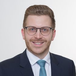 Ein Portrait von Philipp Deuchler. Er hat kurze blonde Haare, trägt eine schwarze Brille und einen dunkelblauen Anzug mit einer hellblauen Krawatte.