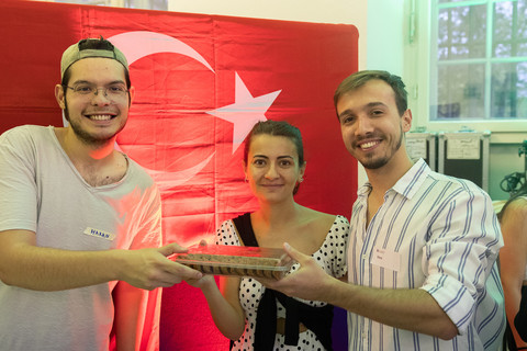 Three people stand smiling in front of a Turkish flag and hold up a Turkish specialty.