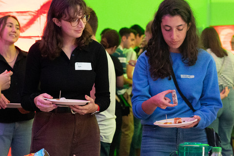 Two students are standing in front of a table with food. Both are holding a plate.